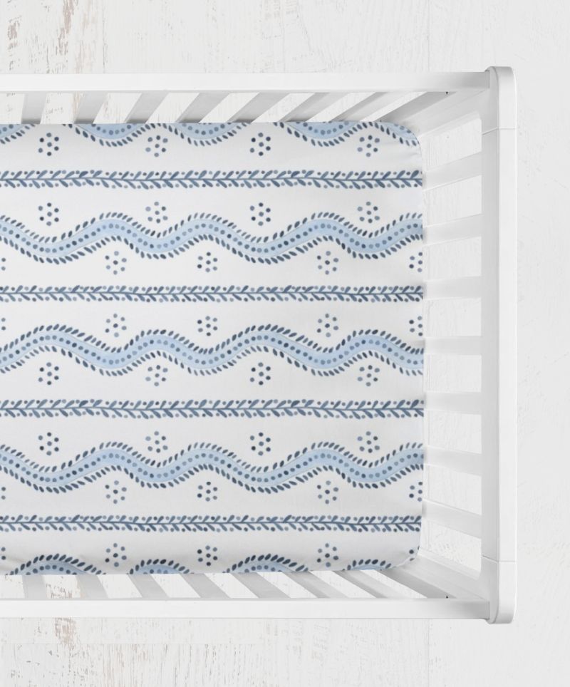 A fitted sheet with a blue and white geometric pattern, shown inside a white wooden crib. Andrew Blue and White Stripes Crib Sheet - The Creative Raccoon 