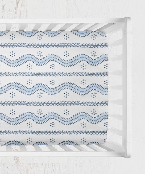 A fitted sheet with a blue and white geometric pattern, shown inside a white wooden crib. Andrew Blue and White Stripes Crib Sheet - The Creative Raccoon 