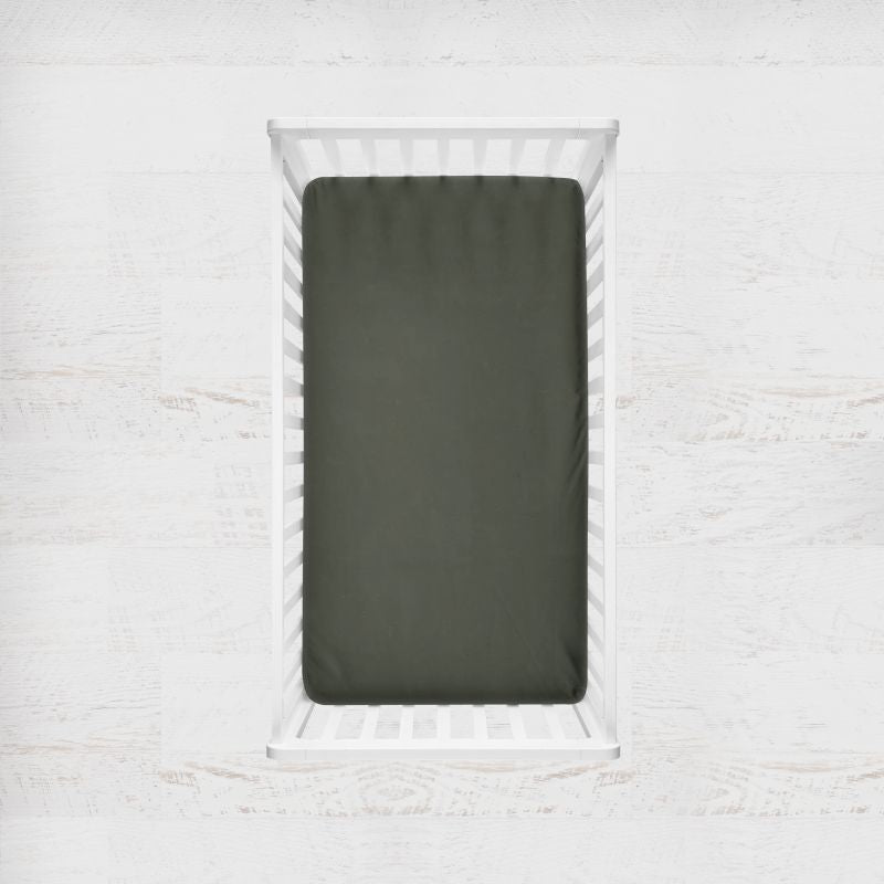 An army green fitted crib sheet shown inside a white wooden crib, set against a light, textured white wall background. Army Green Crib Sheet - The Creative Raccoon