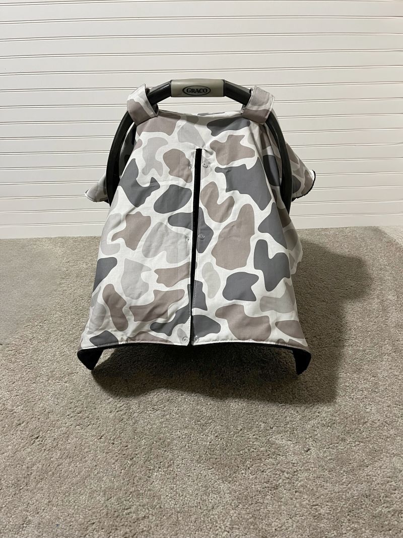 A baby car seat canopy cover with a gray, white, and taupe camouflage pattern, shown on a baby car seat against a white paneled wall and carpeted floor. Baby Car Seat Canopy Cover Camo - The Creative Raccoon