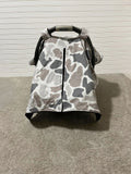 A baby car seat canopy cover with a gray, white, and taupe camouflage pattern, shown on a baby car seat against a white paneled wall and carpeted floor. Baby Car Seat Canopy Cover Camo - The Creative Raccoon