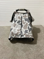 A baby car seat canopy cover with a gray, white, and taupe camouflage pattern, shown on a baby car seat against a white paneled wall and carpeted floor. Baby Car Seat Canopy Cover Camo - The Creative Raccoon