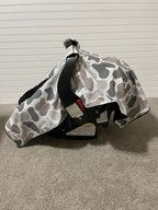 A baby car seat cover with a gray, white, and black camo print pattern draped over a black infant car seat on a carpeted floor against a white paneled wall. Baby Car Seat Canopy Cover Camo - The Creative Raccoon