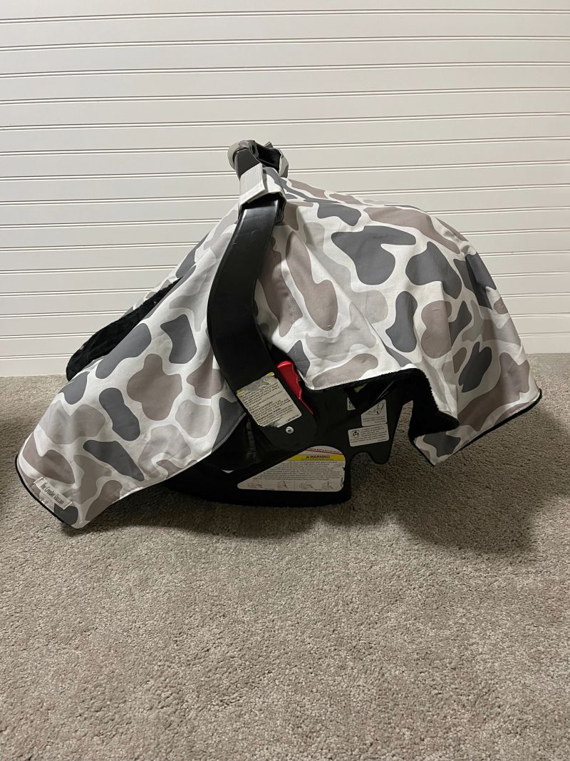 A baby car seat cover with a gray, white, and black camo print pattern draped over a black infant car seat on a carpeted floor against a white paneled wall. Baby Car Seat Canopy Cover Camo - The Creative Raccoon