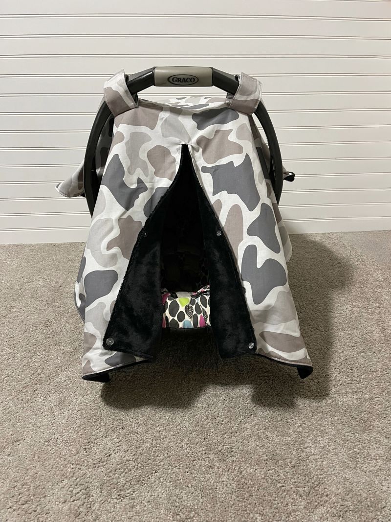 A baby car seat canopy cover with a gray, white, and black camouflage pattern, draped over a black Graco car seat on a beige carpeted floor. Baby Car Seat Canopy Cover Camo - The Creative Raccoon