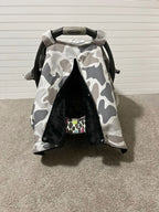 A baby car seat canopy cover with a gray, white, and black camouflage pattern, draped over a black Graco car seat on a beige carpeted floor. Baby Car Seat Canopy Cover Camo - The Creative Raccoon