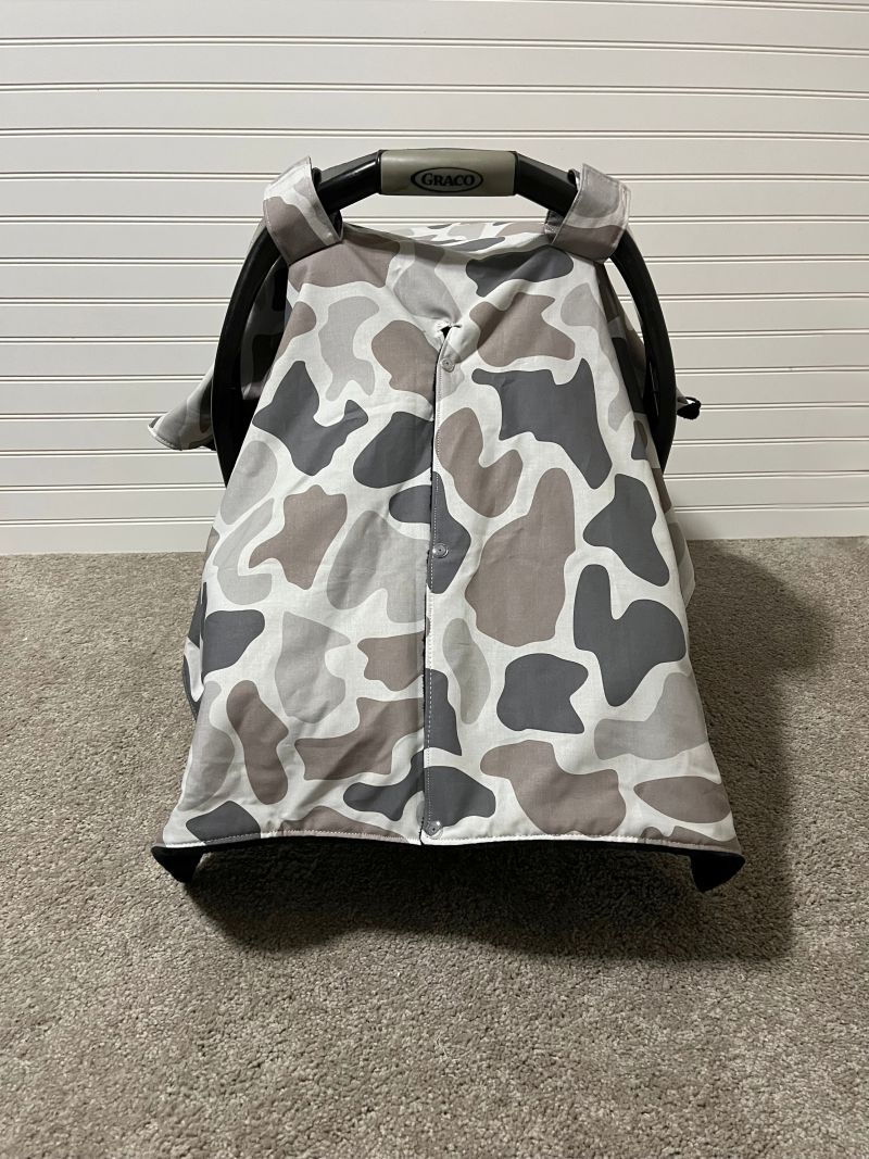 A baby car seat canopy cover with a gray, white, and brown camouflage pattern, draped over a black Graco car seat on a carpeted floor against a white paneled wall. Baby Car Seat Canopy Cover Camo - The Creative Raccoon