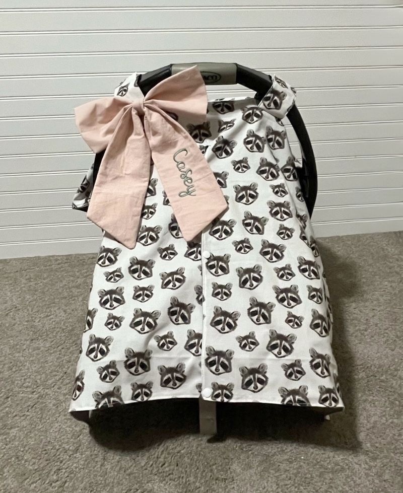A baby car seat cover with a white background and an all-over black raccoon print, featuring a large light pink fabric bow with the word 'cozy' embroidered on it. The product is displayed on a black baby car seat against a white paneled wall and gray carpet. Baby Car Seat Canopy Cover Raccoon with a bow - The Creative Raccoon