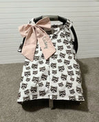 A baby car seat cover with a white background and an all-over black raccoon print, featuring a large light pink fabric bow with the word 'cozy' embroidered on it. The product is displayed on a black baby car seat against a white paneled wall and gray carpet. Baby Car Seat Canopy Cover Raccoon with a bow - The Creative Raccoon