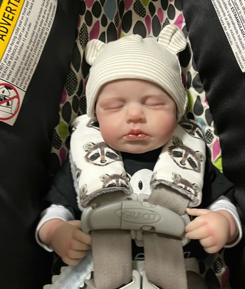 A close-up of a realistic baby doll with closed eyes, wearing a cream ribbed beanie with bear ears and a raccoon-print car seat strap covers, seated in a Graco car seat. Baby Car Seat Strap Covers Raccoon - The Creative Raccoon