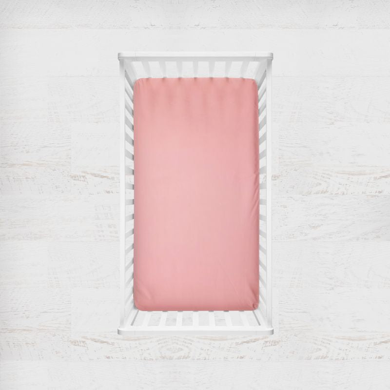 A pink fitted crib sheet displayed inside a white wooden baby crib, shown from a top-down view against a light, textured white background. Baby Pink Crib Sheet - The Creative Raccoon