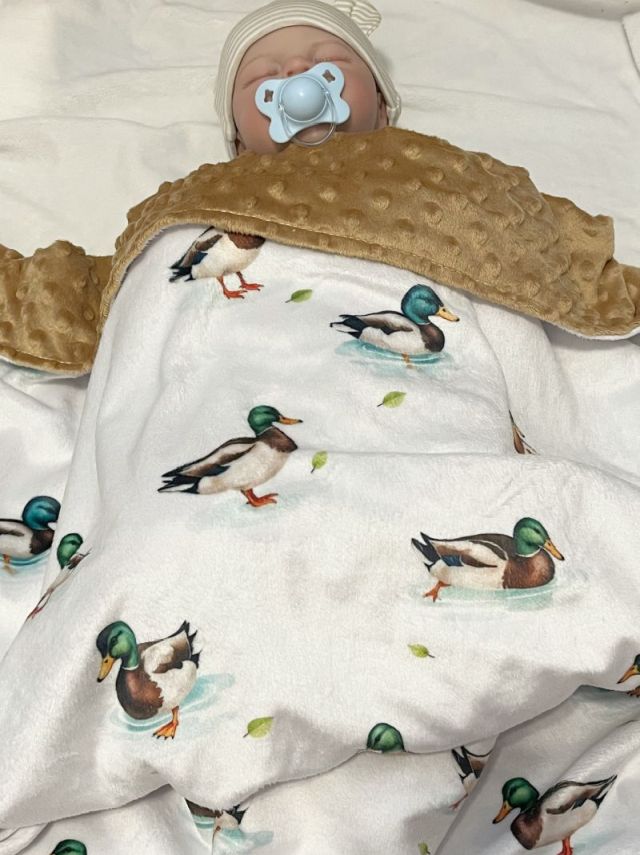 Baby swaddled in a blanket with duck pattern, wearing a beige hat and pacifier. Mallard Duck Baby Blanket - The Creative Racoon