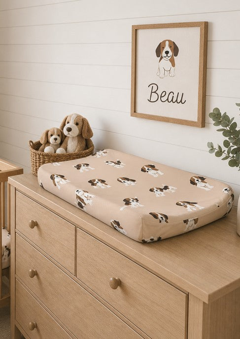 A beige baby changing pad with a brown and white beagle dog print sits on a light wood dresser in a nursery setting. A framed print of a beagle named 'Beau' hangs on the white shiplap wall above, next to a basket of plush dogs.
Beagle Change Pad Cover - The Creative Raccoon