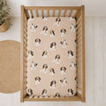 A wooden crib viewed from above, fitted with a beige crib sheet printed with a repeating pattern of brown and white beagle dogs. The crib is placed on a light wood floor next to a woven jute rug.
Beagle Crib Sheet - The Creative Raccoon