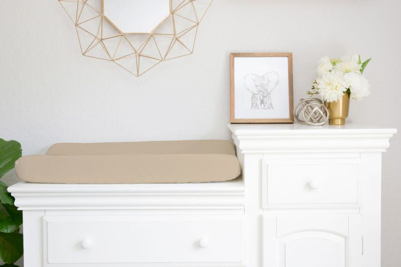 A beige changing pad cushion resting on a white dresser in a bright nursery or nursery-like room, with a framed elephant print, white flowers, and a geometric wall mirror above.
Beige Changing Pad Cover - The Creative Raccoon
