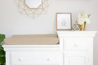 A beige changing pad cushion resting on a white dresser in a bright nursery or nursery-like room, with a framed elephant print, white flowers, and a geometric wall mirror above.
Beige Changing Pad Cover - The Creative Raccoon