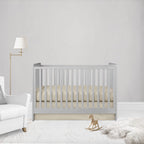 A light gray wooden crib with vertical slats, styled in a minimalist nursery setting with a cream mattress, a white armchair, a gold floor lamp, and a small wooden rocking horse.
Beige Crib Sheet & Flat Crib Skirt - The Creative Raccoon