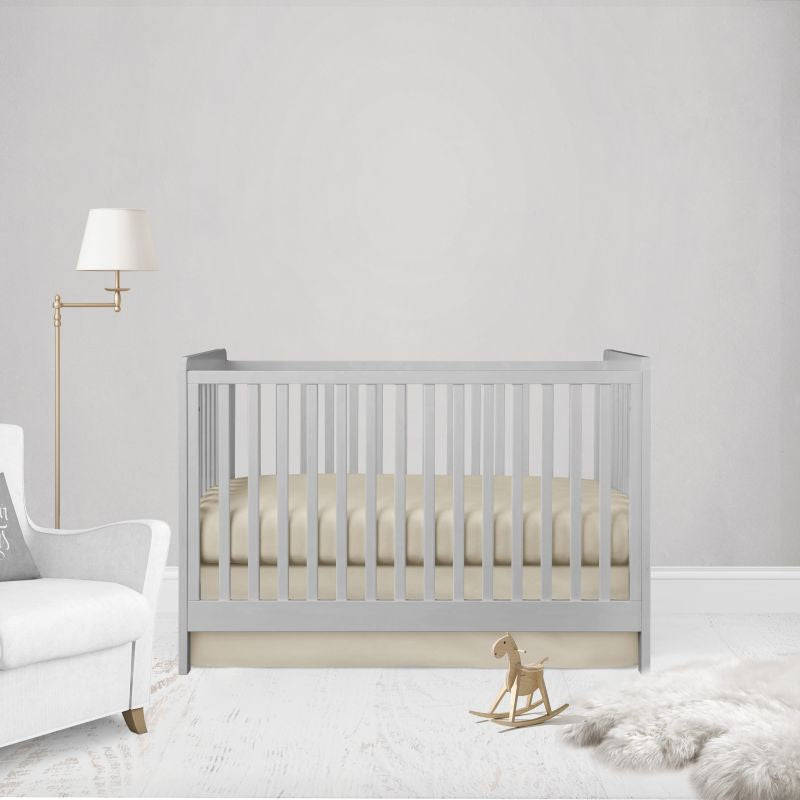 A light gray wooden crib with vertical slats, styled in a minimalist nursery setting with a cream mattress, a white armchair, a gold floor lamp, and a small wooden rocking horse.
Beige Crib Sheet & Flat Crib Skirt - The Creative Raccoon