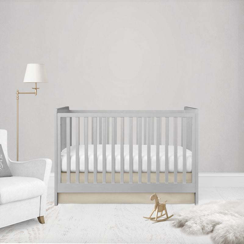 A light gray wooden crib with white bedding, styled in a minimalist nursery setting with a white armchair, a gold floor lamp, a wooden rocking horse, and a white faux fur rug against a light gray wall.
Beige Flat Crib Skirt - The Creative Raccoon