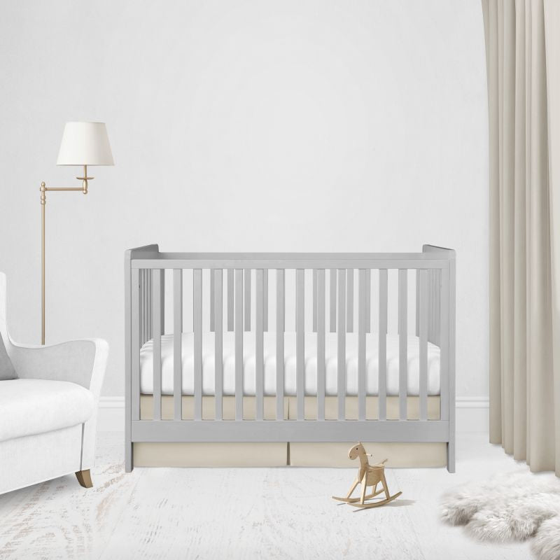 A light gray wooden baby crib with white bedding, styled in a bright, modern nursery with a white armchair, floor lamp, beige curtains, and a small wooden rocking horse.
Beige Pleat Crib Skirt - The Creative Raccoon