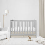A light gray wooden baby crib with white bedding, styled in a bright, modern nursery with a white armchair, floor lamp, beige curtains, and a small wooden rocking horse.
Beige Pleat Crib Skirt - The Creative Raccoon