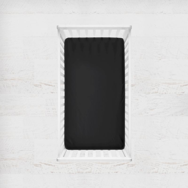 A black fitted crib sheet shown inside a white wooden crib, viewed from directly above against a light textured wall background. Black Crib Sheet - The Creative Raccoon