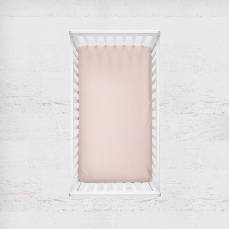A pale blush pink fitted sheet displayed inside a white wooden baby crib, shown from a top-down view against a light, textured white background. Blush Light Pink Crib Sheet - The Creative Raccoon