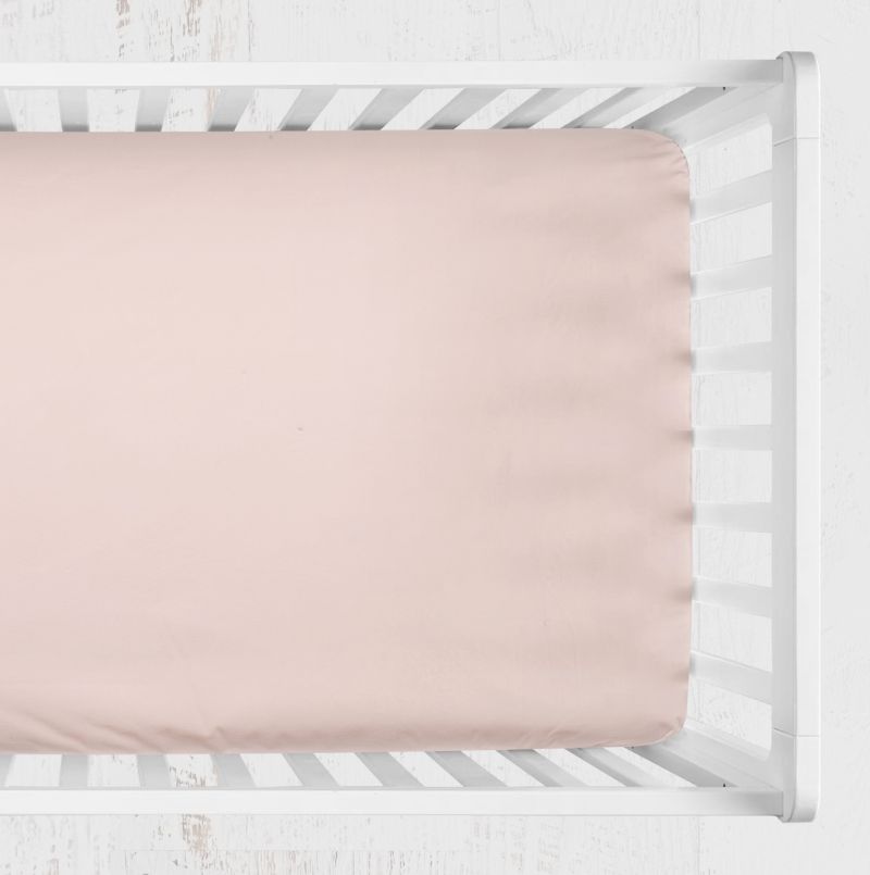 A pale blush pink fitted sheet displayed inside a white wooden baby crib, shown from a top-down view against a light, textured white background. Blush Light Pink Crib Sheet - The Creative Raccoon