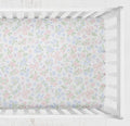 A fitted crib sheet with a delicate pastel floral and bow pattern in pink, blue, and green, shown inside a white wooden crib. Bow Crib Sheet - The Creative Raccoon