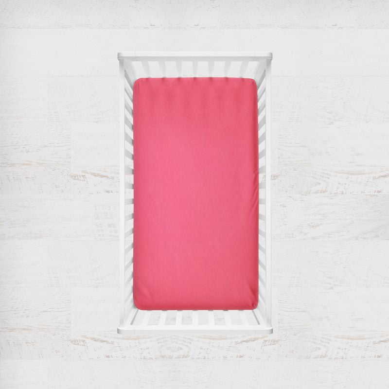 A bright pink fitted sheet on a mattress inside a white wooden baby crib, shown from a top-down view against a white textured wall. Bright Pink Crib Sheet - The Creative Raccoon
