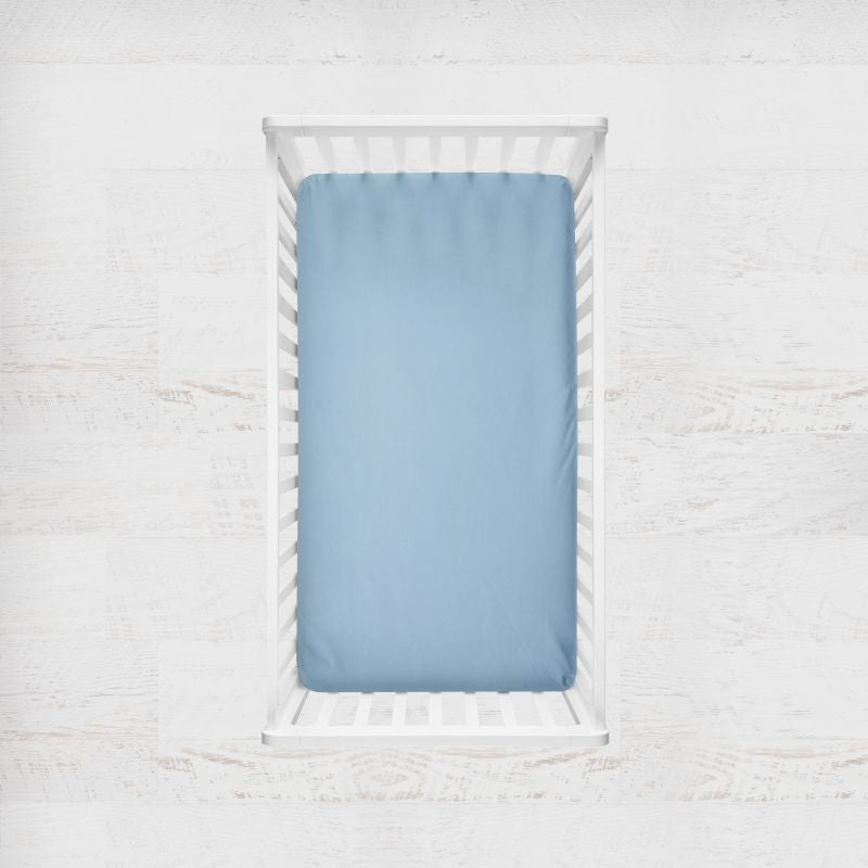 A light blue fitted crib sheet displayed inside a white wooden baby crib, shown from a top-down view against a white textured wall. Candy Blue Crib Sheet - The Creative Raccoon 