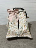 Floral patterned car seat cover with a pink bow and embroidered name on a gray floor. Car Seat Canopy Daisy - The Creative Raccoon