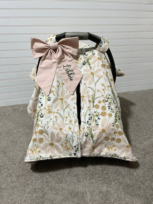 Floral patterned car seat cover with a pink bow and embroidered name on a gray floor. Car Seat Canopy Daisy - The Creative Raccoon