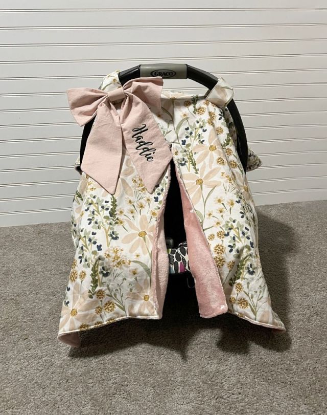 Floral baby car seat cover with pink bow and name on a gray floor. Car Seat Canopy Daisy - The Creative Raccoon