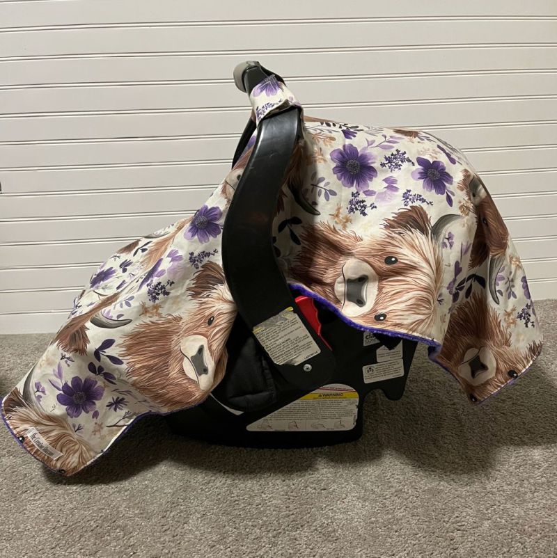 Floral-patterned car seat cover with highland cow design draped over a car seat against a neutral background. Car Seat Canopy Highland Cows - The Creative Raccoon