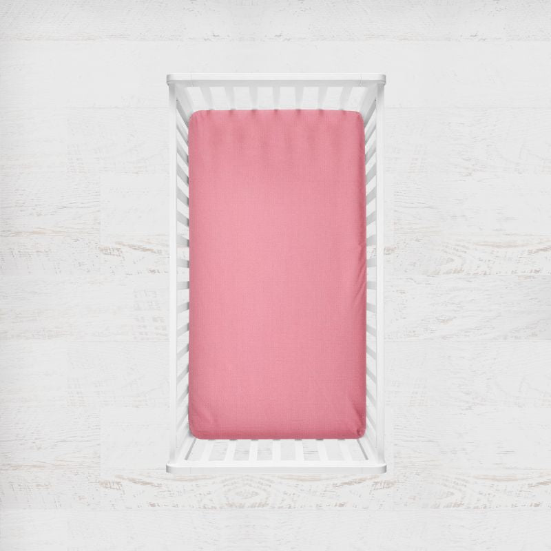 A pink fitted crib sheet shown from above inside a white wooden crib on a light textured white background. Carnation Pink Crib Sheet - The Creative Raccoon