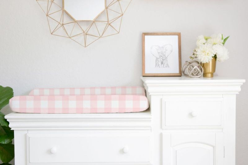 White dresser with pink checkered changing pad, framed picture, and decorative items in a nursery. Changing Pad Cover Light Pink Gingham - The Creative Raccoon
