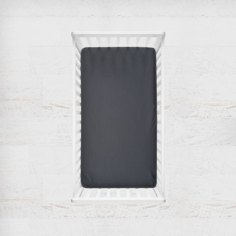 A dark charcoal gray fitted sheet displayed on a white wooden crib, shown from a top-down perspective against a light, textured white wall. Charcoal Gray Crib Sheet - The Creative Raccoon