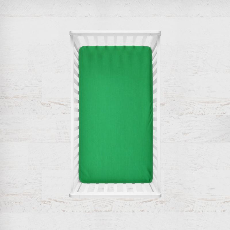 A bright green fitted crib sheet inside a white wooden crib, shown from a top-down view against a light textured white wall. Clover Green Crib Sheet - The Creative Raccoon