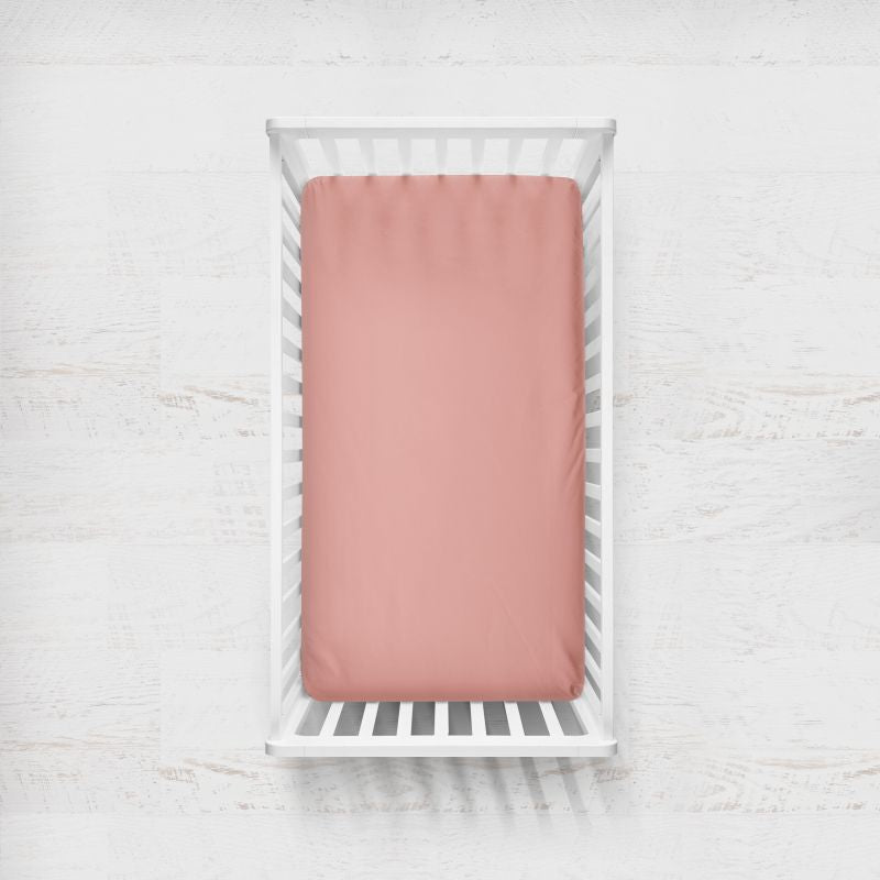 A dusty coral pink fitted crib sheet displayed inside a white wooden baby crib, viewed from a top-down perspective against a white textured wall. Coral Pink Crib Sheet - The Creative Raccoon