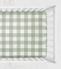 A green and white gingham check crib sheet fitted inside a white wooden crib, viewed from directly above. 
Crib Sheet - Sage Gingham Check - The Creative Raccoon