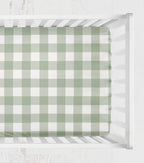A green and white gingham check crib sheet fitted inside a white wooden crib, viewed from directly above. 
Crib Sheet - Sage Gingham Check - The Creative Raccoon