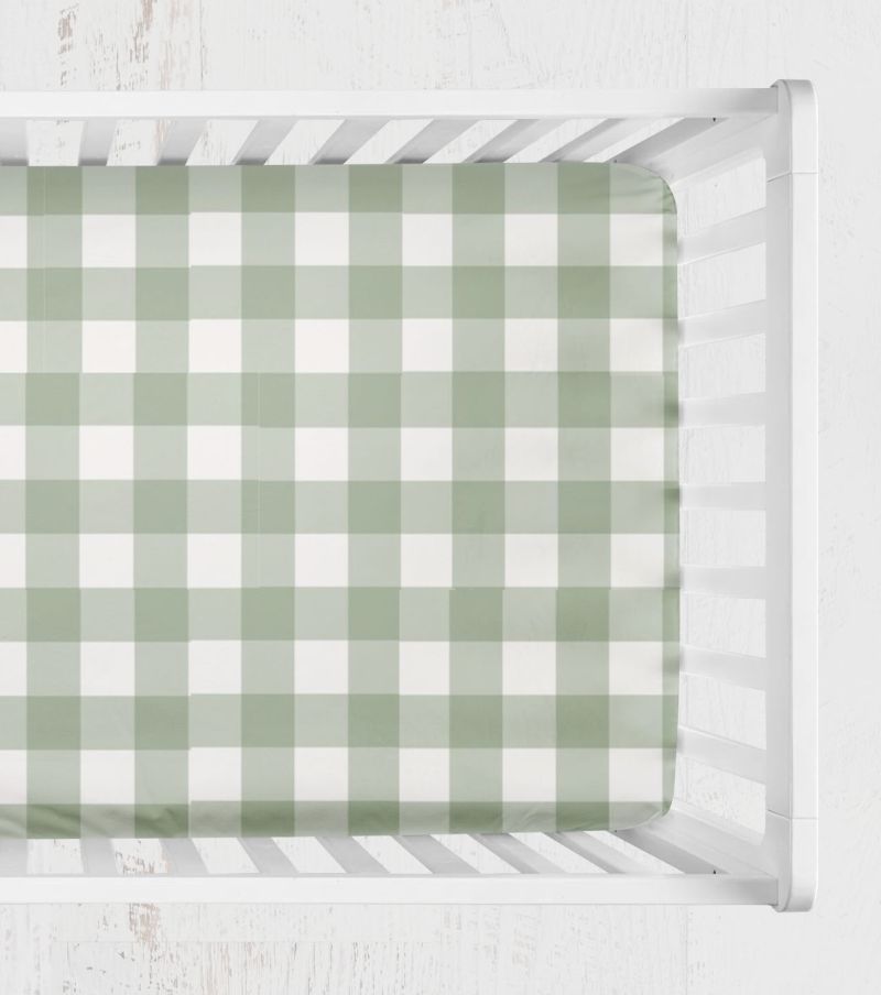 A green and white gingham check crib sheet fitted inside a white wooden crib, viewed from directly above. 
Crib Sheet - Sage Gingham Check - The Creative Raccoon