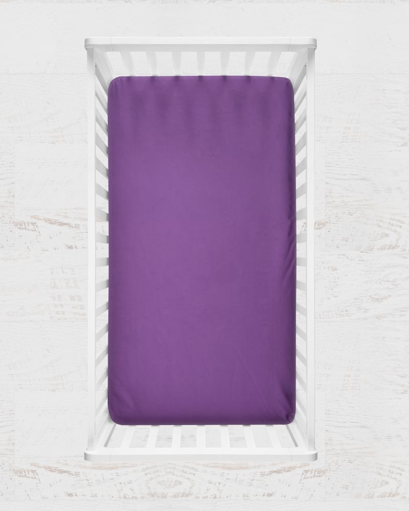 A solid purple fitted crib sheet shown inside a white wooden crib, set against a light, textured white background. Crocus Purple Crib Sheet - The Creative Raccoon