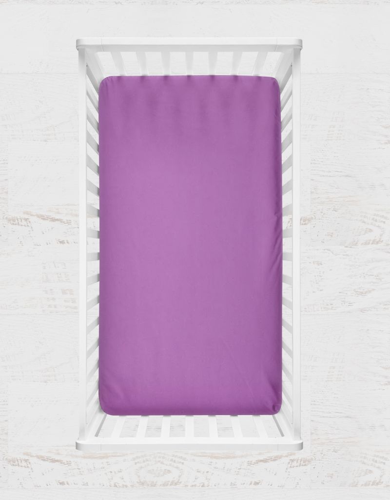 A solid purple fitted crib sheet shown inside a white wooden crib, set against a light, textured white background. Dahlia Purple Crib Sheet - The Creative Raccoon