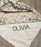 Floral-patterned blanket with 'OLIVIA' embroidery on a beige surface. Daisy Baby Blanket - The Creative Raccoon