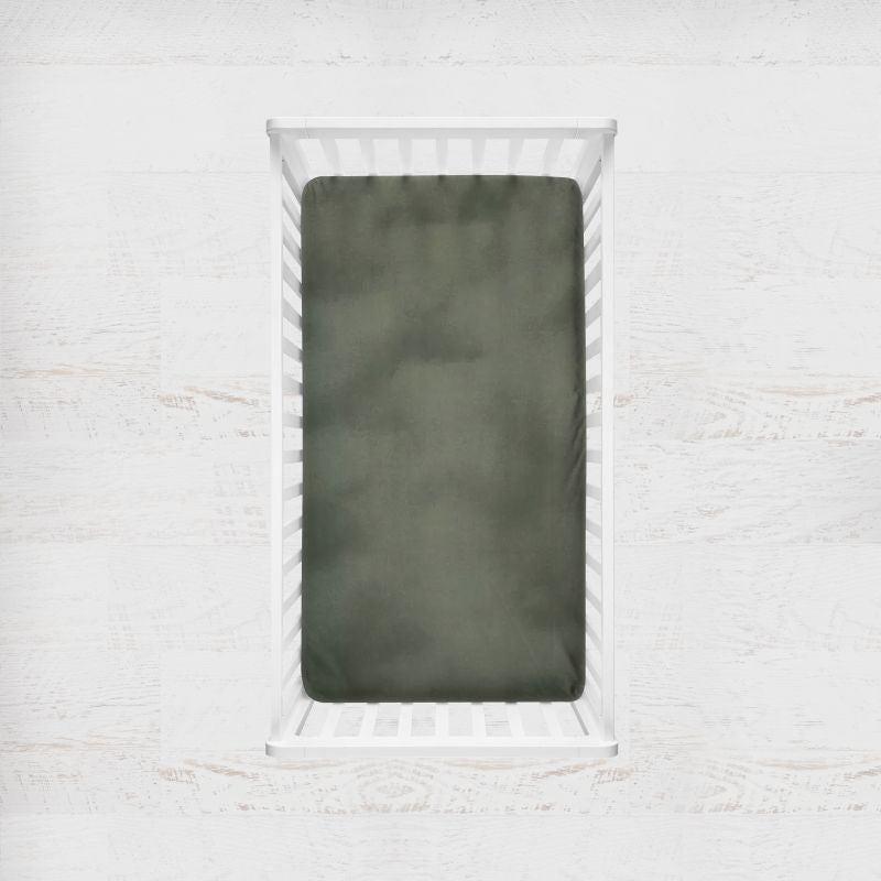 An olive green fitted crib sheet shown inside a white wooden baby crib, set against a light, textured white wall background. Dark Sage Green Crib Sheet - The Creative Raccoon