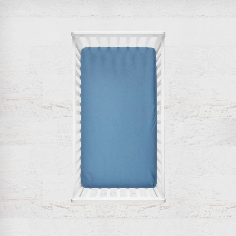 A delft blue fitted crib sheet on a mattress inside a white wooden crib, shown from a top-down view against a white textured wall. Delft Blue Crib Sheet - The Creative Raccoon
