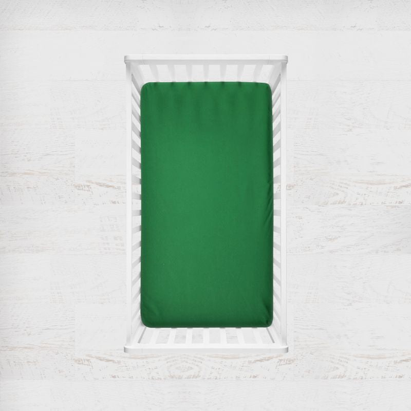 A bright forest green fitted crib sheet shown inside a white wooden crib, photographed from above against a light textured wall. Forest Green Crib Sheet - The Creative Raccoon