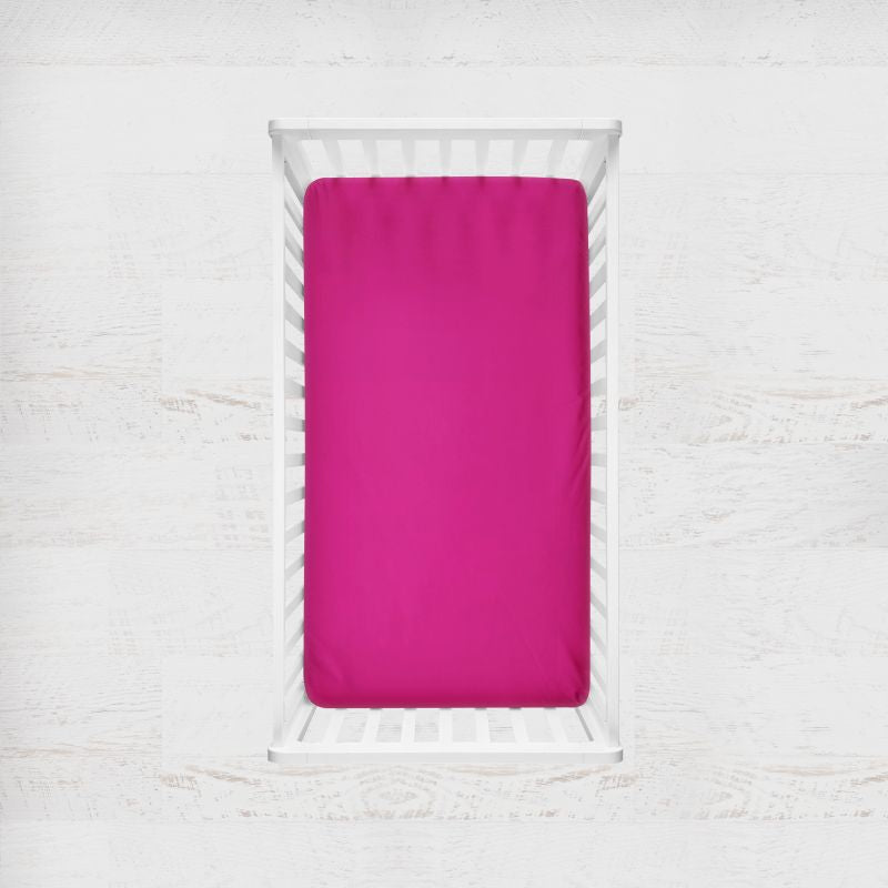 A bright fuchsia pink fitted crib sheet inside a white wooden baby crib, shown from a top-down view against a white textured background. Fushia Bright Pink Crib Sheet - The Creative Raccoon